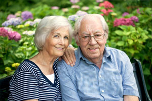 A couple sitting in their garden and smiling at the camera. You can see flowers behind them.