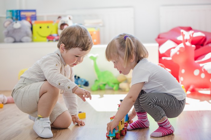 A young boy and girl playing with a toy train on the nusery floor. Soft toys are scattered around them.