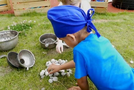 Two children are picking up pebbles outside and placing them into old cooking pots.